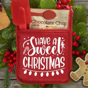 Op de afbeelding: Een rode ovenwant met de tekst "Have a Sweet Christmas" in witte letters. De ovenwant heeft een zakje met een pakje chocolade-chip koekjesmix en een rode spatel.