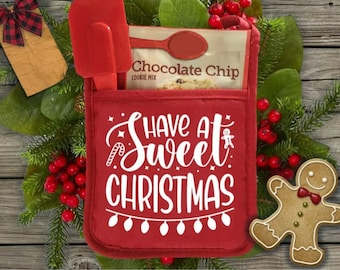 Op de afbeelding: Een rode ovenwant met de tekst "Have a Sweet Christmas" in witte letters. De ovenwant heeft een zakje met een pakje chocolade-chip koekjesmix en een rode spatel.