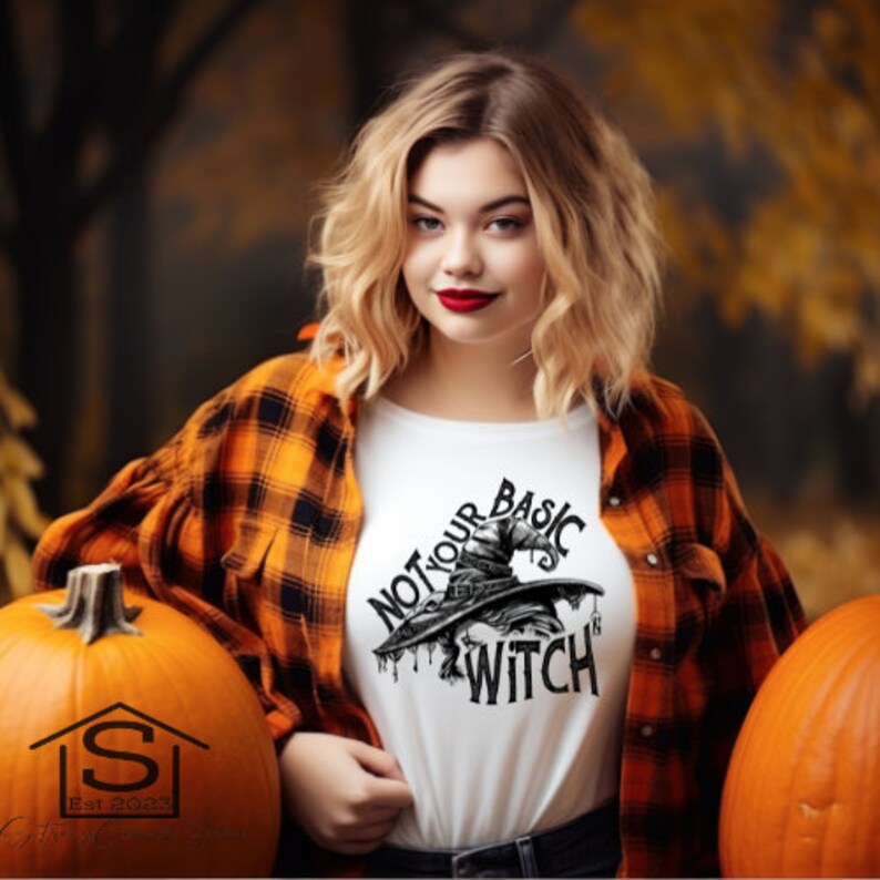 Może przedstawiać: Biała koszulka z napisem "NOT YOUR BASIC WITCH" i grafiką kapelusza czarownicy. Osoba ma na sobie pomarańczowo-czarną koszulę w kratę i jest otoczona dyniami, idealna na Halloween.