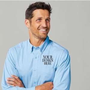 May include: Light blue button-down shirt with the text "YOUR DESIGN HERE" printed on the chest. The shirt has a button-down collar and a chest pocket. The person is wearing gray pants.