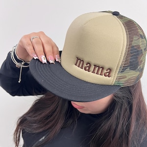 May include: A beige and camouflage trucker hat with the word "mama" embroidered in brown. The hat has a black brim and a mesh back. The hat is a stylish accessory.