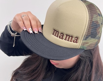 May include: A beige and camouflage trucker hat with the word "mama" embroidered in brown. The hat has a black brim and a mesh back. The hat is a stylish accessory.