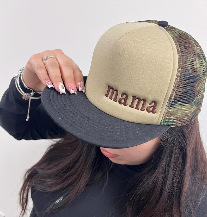 May include: A beige and camouflage trucker hat with the word "mama" embroidered in brown. The hat has a black brim and a mesh back. The hat is a stylish accessory.