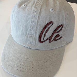 May include: A light gray baseball cap with a structured crown and a curved brim. The front features a dark red script logo that reads Cle, with the letter C designed to resemble a football, complete with a textured surface and white stitching.