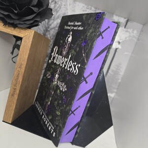 May include: A paperback book with a black cover and purple spine. The cover features a sword and the title "Powerless" in white lettering. The author's name, Lauren Roberts, is printed in white on the spine.