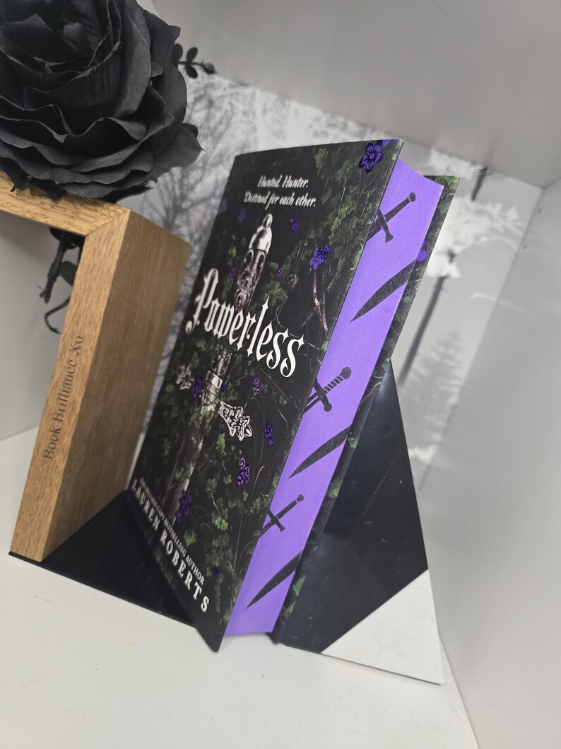 May include: A paperback book with a black cover and purple spine. The cover features a sword and the title "Powerless" in white lettering. The author's name, Lauren Roberts, is printed in white on the spine.