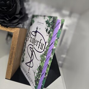 May include: A paperback book titled "Powerful" by  S.J. Roberts with a purple bookmark. The cover features a white background with a black snake wrapped around a sword and green leaves.