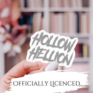 Könnte beinhalten: Ein grauer Aufkleber mit den Worten "Hollow Hellion" in einer stilisierten Schriftart. Der Aufkleber wird in einer Hand mit weißem Nagellack gehalten. Der Text "OFFICIALLY LICENSED" steht darunter.