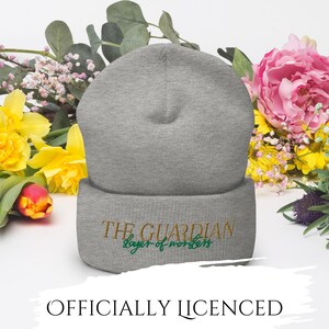 May include: A gray beanie with the embroidered text "THE GUARDIAN slayer of monsters" in gold and green. The hat is displayed in front of a floral arrangement with yellow, pink, and white flowers. The text "OFFICIALLY LICENSED" is at the bottom.