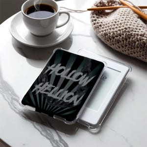 Peut inclure: Une coque de protection transparente pour liseuse électronique avec le texte "Hollow Hellion" en gris. Le motif présente un fond noir et gris avec un motif de rayons de soleil et des détails de chaînes. Une liseuse blanche est visible.