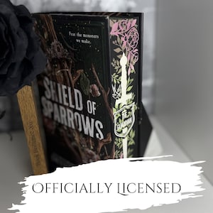 Op de afbeelding: Een hardcover boek getiteld "Shield of Sparrows" met een donkere omslag met een takontwerp en de tekst "Fear the monsters we make." Het boek heeft een decoratieve rug met bloemaccenten en de tekst "The Guardian." De tekst "Officially Licensed" staat onderaan.