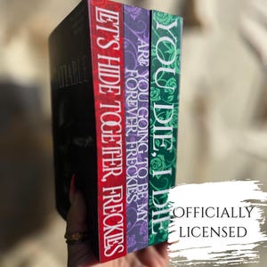 May include: Three books with vibrant spines are stacked together. The first book is black with the title "Satiable". The second book is red with the text "Let's Hide Together, Freckles". The third book is purple with the text "Are You Going To Be My Forever Freckles". The fourth book is green with the text "You Die, I Die".