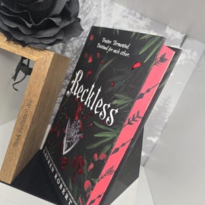 May include: A black book with a red spine and the title "Reckless" in white lettering. The book is standing upright on a black surface. The text on the cover reads "Traitor. Tormented. Destined for each other."