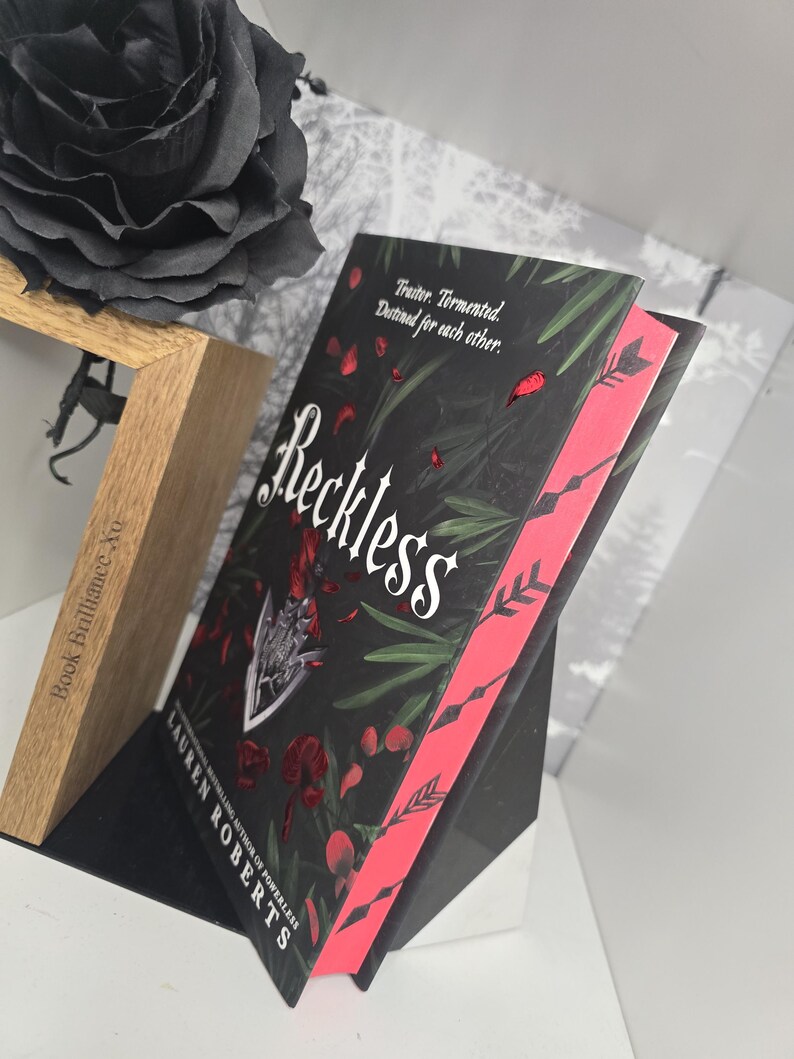 May include: A black book with a red spine and the title "Reckless" in white lettering. The book is standing upright on a black surface. The text on the cover reads "Traitor. Tormented. Destined for each other."