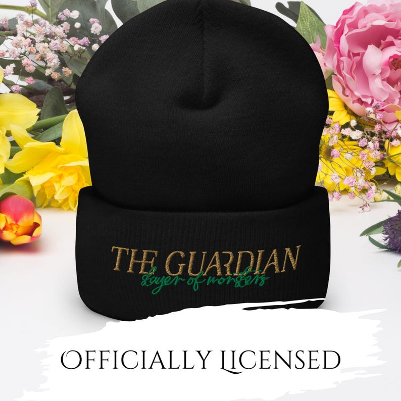 May include: Black beanie with the text "THE GUARDIAN" embroidered in gold and "slayer of monsters" in green. The beanie is a winter accessory, likely made of a soft, warm material. The words "OFFICIALLY LICENSED" are printed below.