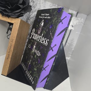 May include: A paperback book titled "Powerless" by Lauren Roberts, a bestselling author. The book cover features a dark green background with a sword and a tower, and the title is written in white with a purple outline. The book is on a black and wooden display stand.