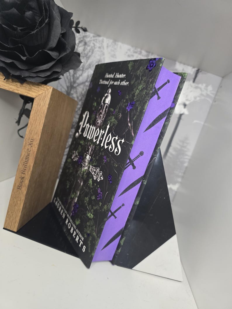 May include: A paperback book titled "Powerless" by Lauren Roberts, a bestselling author. The book cover features a dark green background with a sword and a tower, and the title is written in white with a purple outline. The book is on a black and wooden display stand.