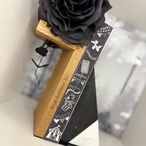 Puede incluir: Un soporte de libro decorativo de madera con un diseño en blanco y negro, que sostiene un libro con la palabra "ODDITY" e ilustraciones con temática de carnaval. Una gran rosa negra se encuentra encima del soporte. El soporte tiene el texto "Book Brilliance Xo".