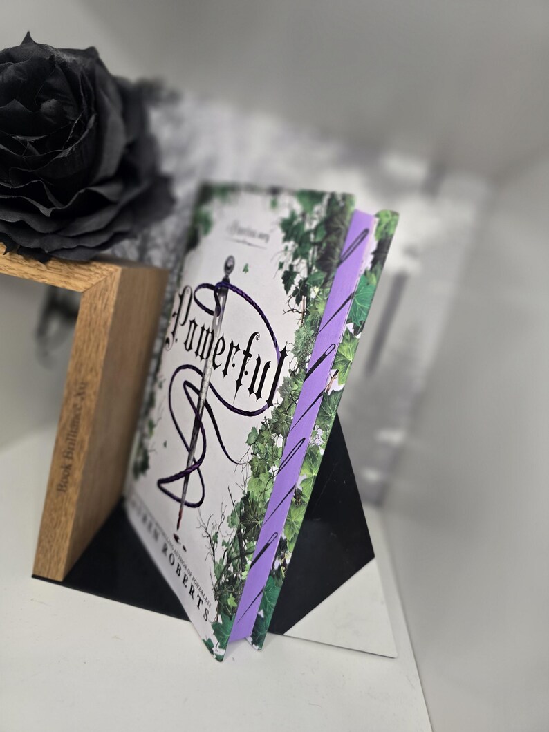 May include: A paperback book titled "Powerful" by  S.J. Roberts with a purple bookmark. The cover features a white background with a black snake wrapped around a sword and green leaves.