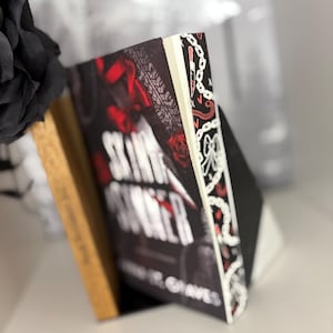 Può includere: Un libro intitolato "Skin Runner" con una copertina nera con rose rosse e impronte di pneumatici. Il dorso è nero con elementi decorativi bianchi. Sullo sfondo è visibile un fiore di stoffa nero.