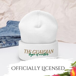 May include: A white beanie with the text "THE GUARDIAN slayer of monsters" embroidered in brown and green. The words "OFFICIALLY LICENSED" are at the bottom of the image. The beanie is made of a soft material.