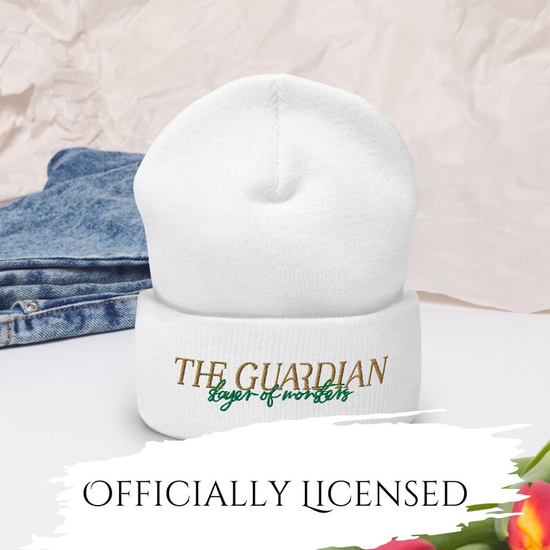 May include: A white beanie with the text "THE GUARDIAN slayer of monsters" embroidered in brown and green. The words "OFFICIALLY LICENSED" are at the bottom of the image. The beanie is made of a soft material.