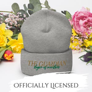 May include: A gray beanie with the embroidered text "THE GUARDIAN slayer of monsters" in gold and green. The words "OFFICIALLY LICENSED" are printed below. The beanie is surrounded by colorful flowers.