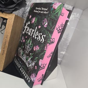 May include: A paperback book titled "Fearless" by Lauren Roberts, the author of "Powerless". The cover features a dark background with pink roses and green leaves. The spine is pink with black crowns.