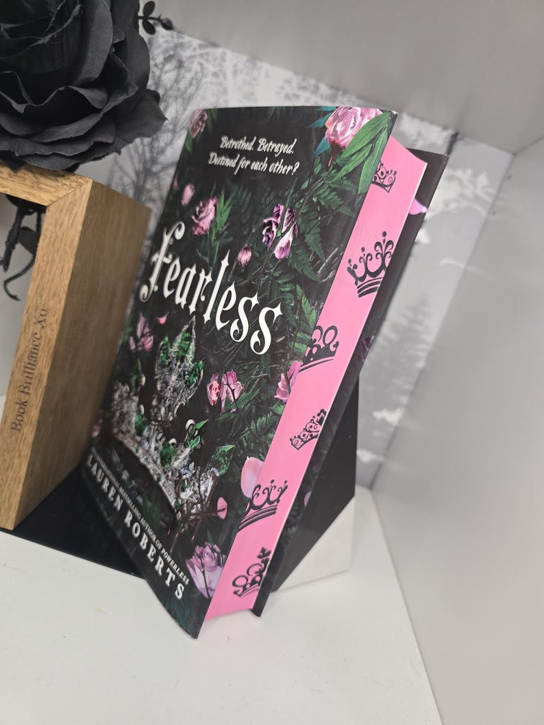 May include: A paperback book titled "Fearless" by Lauren Roberts, the author of "Powerless". The cover features a dark background with pink roses and green leaves. The spine is pink with black crowns.