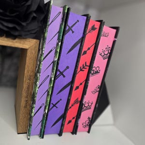 Può includere: Cinque libri rilegati con diversi design. I libri sono impilati uno sopra l'altro. Il libro in basso ha un design floreale verde e nero con un motivo di ago e filo. Il libro successivo è viola con un motivo di spada. Il libro successivo è rosso con un motivo di freccia. Il libro successivo è rosa con un motivo di corona. Il libro in alto è rosa con un motivo di corona. I libri sono tutti intitolati "Book Brilliance XO".