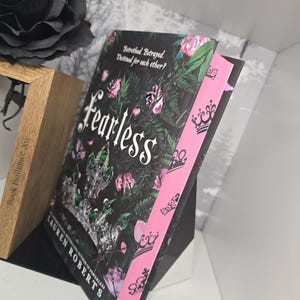May include: A paperback book titled "Fearless" by Lauren Roberts, the book is a sequel to "Powerless". The cover features a black background with a floral design and a crown. The spine is pink with a repeating crown design.
