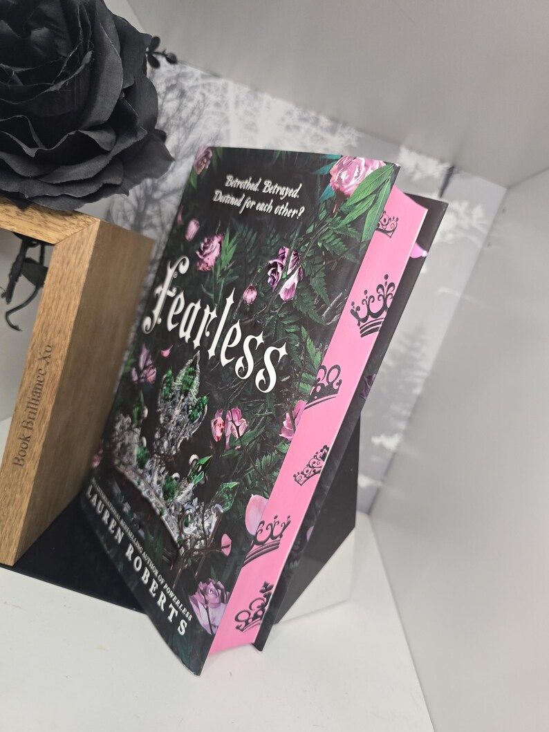 May include: A paperback book titled "Fearless" by Lauren Roberts, the book is a sequel to "Powerless". The cover features a black background with a floral design and a crown. The spine is pink with a repeating crown design.
