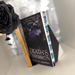 Può includere: Un libro intitolato "Death's Obsession" con una copertina scura raffigurante un corvo. Il libro è appoggiato a un leggio nero. Il dorso ha un bordo bianco con accenti floreali blu. Una rosa nera è sullo sfondo.