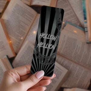 Könnte beinhalten: Ein schwarz-grauer Lesezeichen mit dem Text "Hollow Hellion" in Weiß. Das Lesezeichen hat ein Sonnenstrahl-Design und eine Ketten-Grafik. Der Hintergrund ist eine Sammlung offener Bücher.