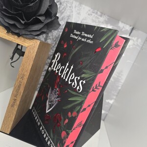 May include: A black book with a red spine and a black rose on a wooden stand. The book cover features a dark green background with red petals and the title "Reckless" in white lettering. The author's name, "Lauren Roberts", is printed in white lettering below the title.