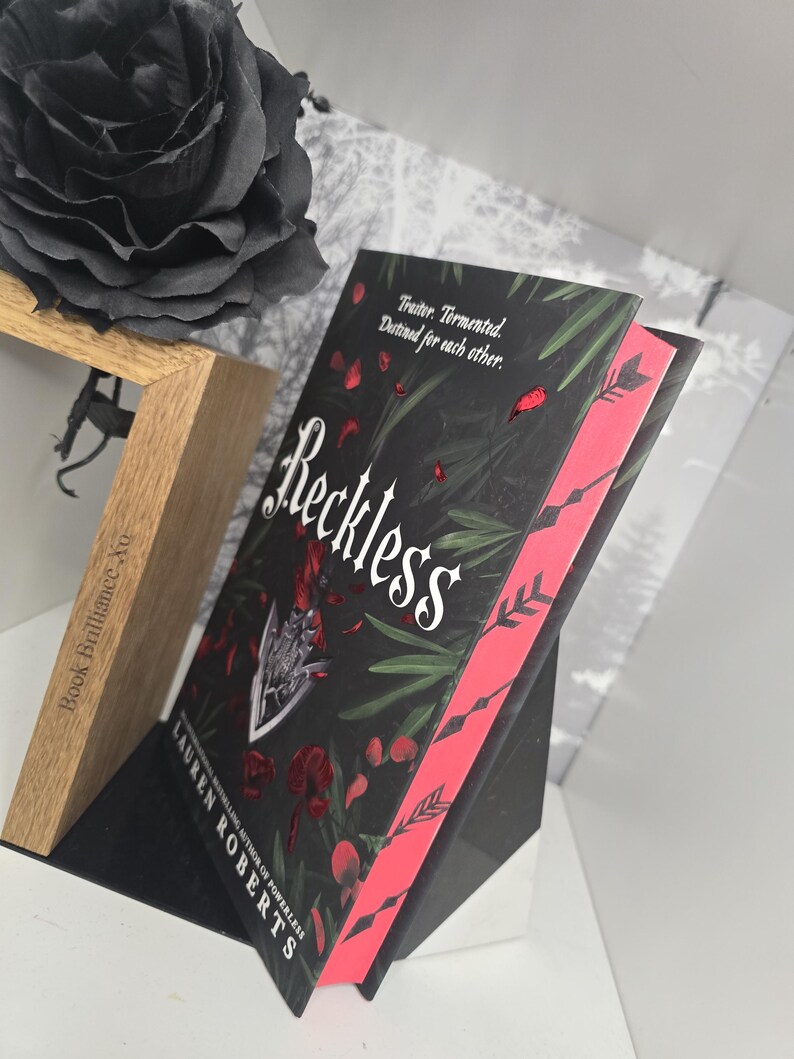 May include: A black book with a red spine and a black rose on a wooden stand. The book cover features a dark green background with red petals and the title "Reckless" in white lettering. The author's name, "Lauren Roberts", is printed in white lettering below the title.