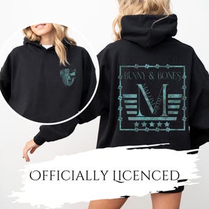 Op de afbeelding: Een zwarte hoodie met een blauwgroene afbeelding op de linkerborst van een schedel met een vlagpatroon. De achterkant toont een groot blauwgroen vierkant kader met de tekst BUNNY & BONES, een grote letter M en vijf sterren.