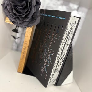 May include: A black book with iridescent lettering and a butterfly design, propped up by a wooden book stand. A large, dark gray rose is positioned above the book. The book is a New York Times Bestseller.