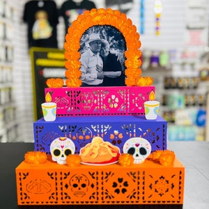 Puede incluir: Un altar de tres niveles, una ofrenda tradicional mexicana del D&iacute;a de Muertos, decorado con cempas&uacute;chil naranja, recortes de papel morado y rosa, y calaveras de az&uacute;car. Una foto enmarcada de una pareja mayor est&aacute; en el nivel superior.