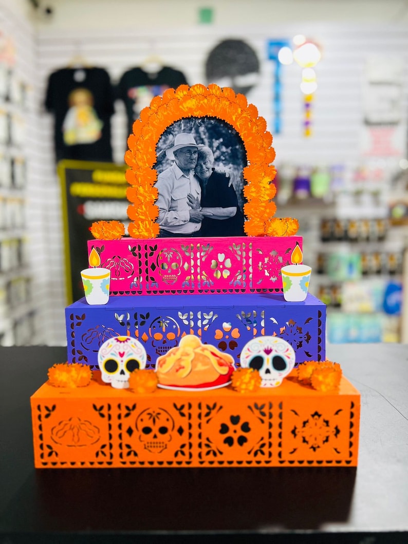 Puede incluir: Un altar de tres niveles, una ofrenda tradicional mexicana del D&iacute;a de Muertos, decorado con cempas&uacute;chil naranja, recortes de papel morado y rosa, y calaveras de az&uacute;car. Una foto enmarcada de una pareja mayor est&aacute; en el nivel superior.