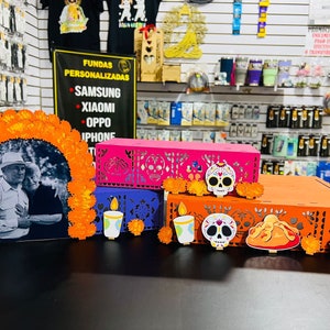 Puede incluir: Un altar de D&iacute;a de Muertos con una foto de una pareja, flores de cempas&uacute;chil naranja, y tres cajas decoradas con calaveras de az&uacute;car, velas y un pan de muerto. Las cajas son rosa, morado y naranja.