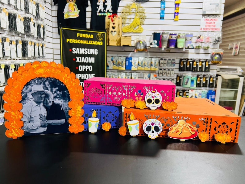 Puede incluir: Un altar de D&iacute;a de Muertos con una foto de una pareja, flores de cempas&uacute;chil naranja, y tres cajas decoradas con calaveras de az&uacute;car, velas y un pan de muerto. Las cajas son rosa, morado y naranja.
