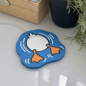 Op de afbeelding: Een blauwe draadloze telefoonoplader met een cartoon eend ontwerp. De oplader heeft een wit eendenlichaam en oranje voeten, met een blauwe waterachtergrond. Andere items zijn boeken, een kaars en een potplant.