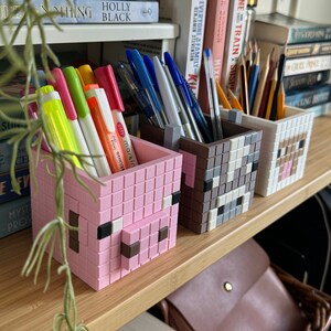 May include: Three Minecraft-themed pencil holders with pixelated animal faces, in pink, grey and brown. The holders are on a wooden shelf with books and a brown leather bag.