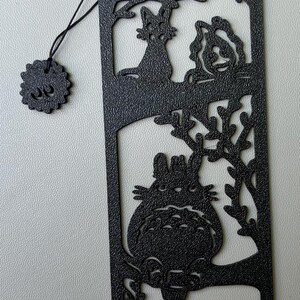 Może przedstawiać: Czarna metalowa zakładka do książki z wyciętym wzorem postaci Totoro z filmu M&oacute;j sąsiad Totoro. Zakładka zawiera r&oacute;wnież dwa koty siedzące pod drzewem.