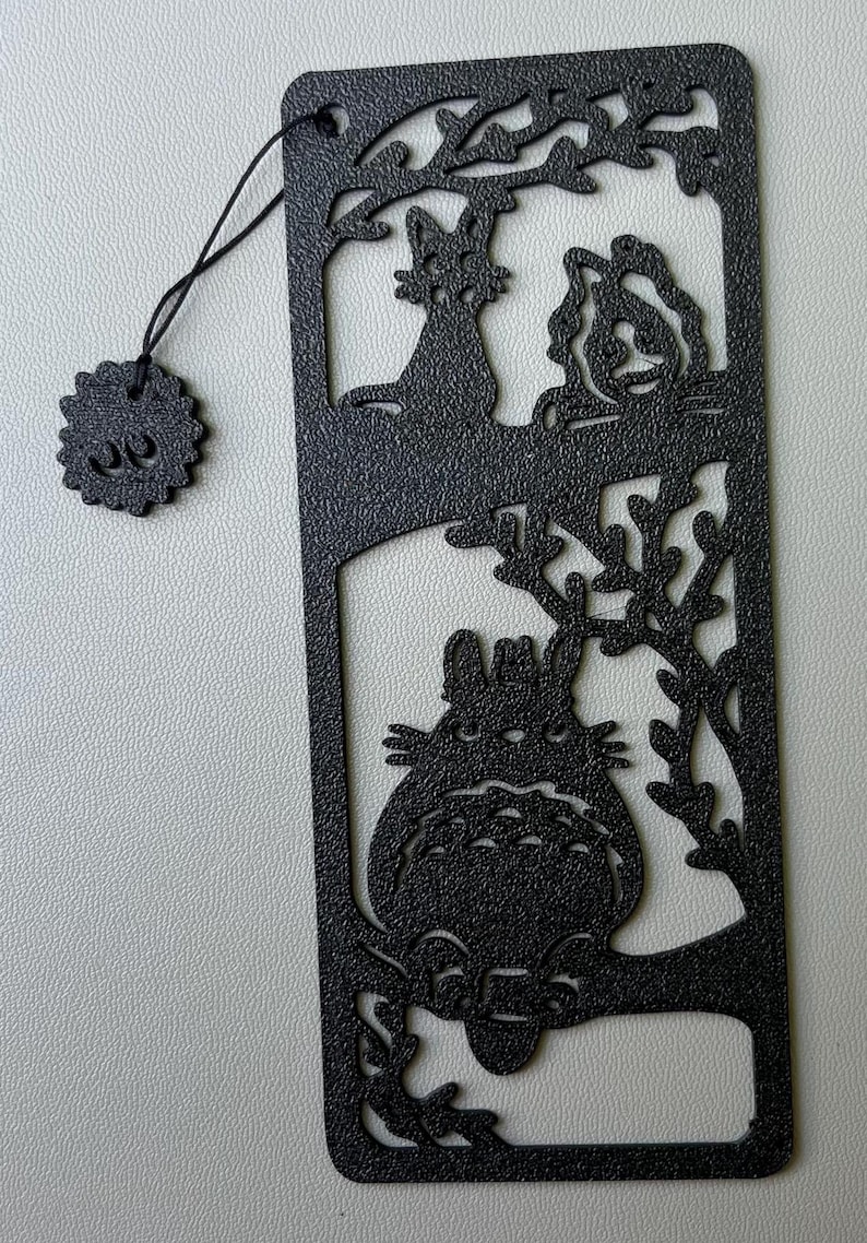 Może przedstawiać: Czarna metalowa zakładka do książki z wyciętym wzorem postaci Totoro z filmu M&oacute;j sąsiad Totoro. Zakładka zawiera r&oacute;wnież dwa koty siedzące pod drzewem.