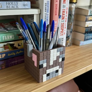 May include: A brown and white pixelated cow-shaped pencil holder with a pink nose and black eyes. The holder is filled with pens and pencils.