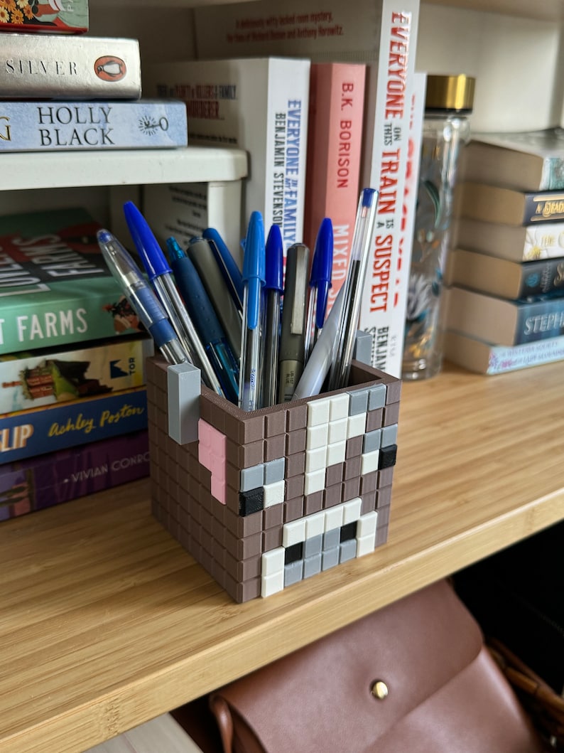 May include: A brown and white pixelated cow-shaped pencil holder with a pink nose and black eyes. The holder is filled with pens and pencils.
