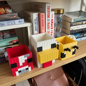 Pode incluir: Tr&ecirc;s pequenos vasos em forma de cubo branco com designs pixelados. Os vasos s&atilde;o vermelhos, amarelos e brancos. Os vasos est&atilde;o em uma prateleira de madeira com livros e outros itens.