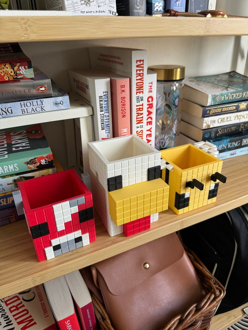 Pode incluir: Tr&ecirc;s pequenos vasos em forma de cubo branco com designs pixelados. Os vasos s&atilde;o vermelhos, amarelos e brancos. Os vasos est&atilde;o em uma prateleira de madeira com livros e outros itens.